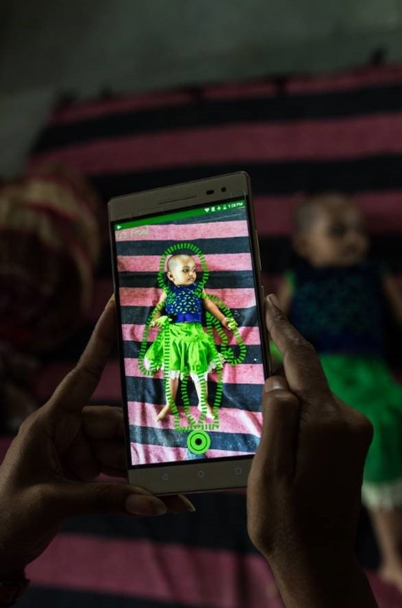 A field worker scans a child with an app-enabled smartphone as the child rests on her back atop a blanket.