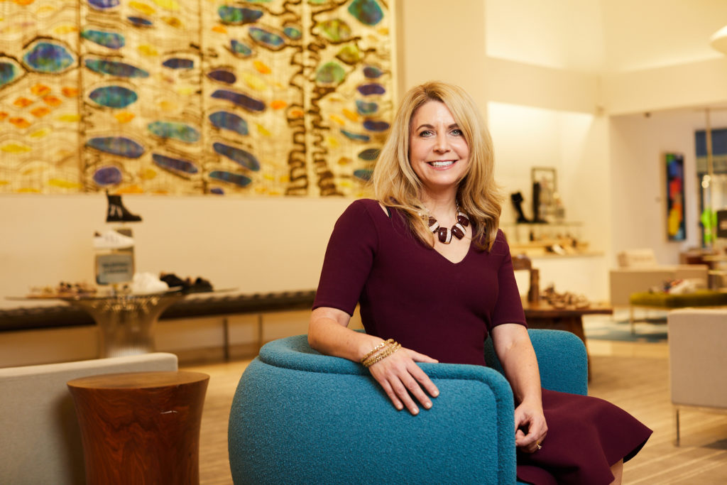Sarah Miller, senior vice president of Neiman Marcus, sits on a blue chair in a maroon dress, smiling.
