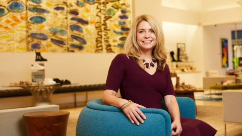 Sarah Miller, senior vice president of Neiman Marcus, sits on a blue chair in a maroon dress, smiling.