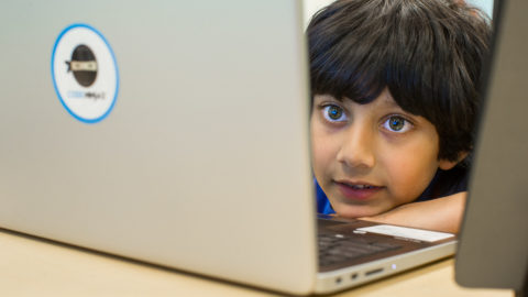 A young boy peers into his laptop with his head perched on the desk.