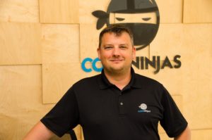 David Graham, founder of Code Ninjas, smiles while standing in front of the organization's logo. 