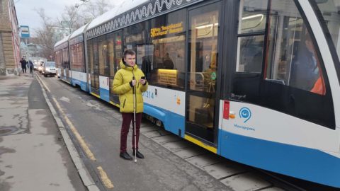 Alexandr stands next to a streetcar with his phone and cane