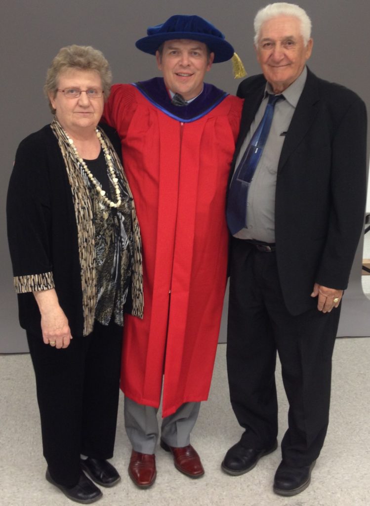 Theo stands between his parents Donna and Wally at a ceremony where he received an honorary doctorate degree. Theo is wearing a red graduation gown and cap. 