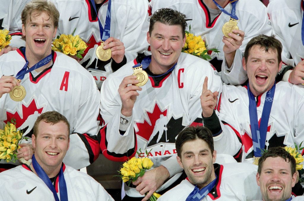 Theo Fleury appears with five other hockey players, all smiling and wearing gold medals, at the 2002 Winter Olympics.