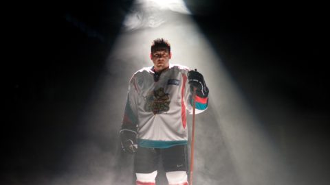 Theo Fleury stands in his skates and hockey uniform on ice under a spotlight.