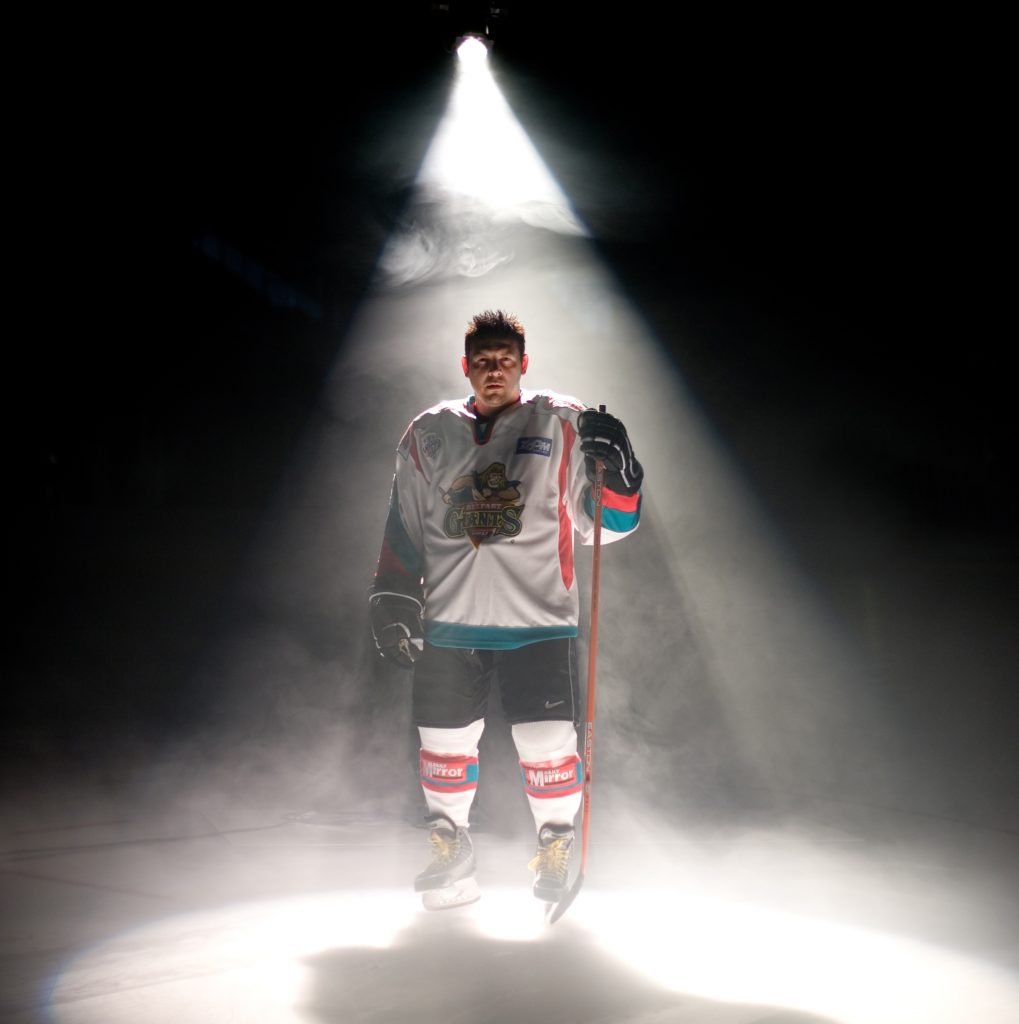 Theo Fleury stands in his skates and hockey uniform on ice under a spotlight.