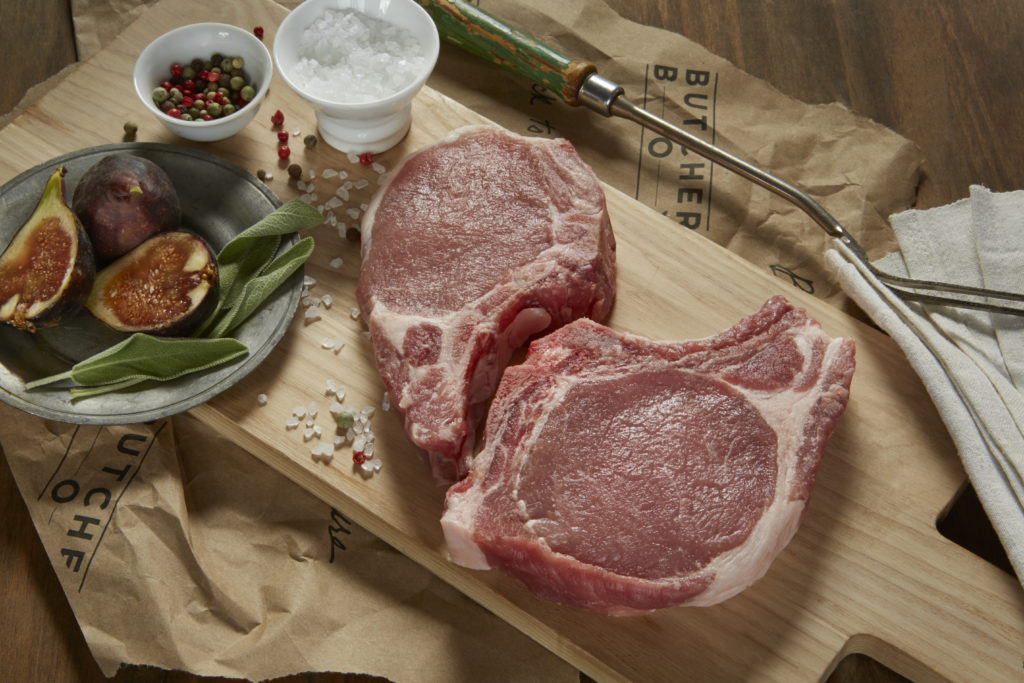 Photo of two bone-in pork chops on a wooden board, with bows of salt and peppercorns and a plate with fresh figs and fresh sage leaves