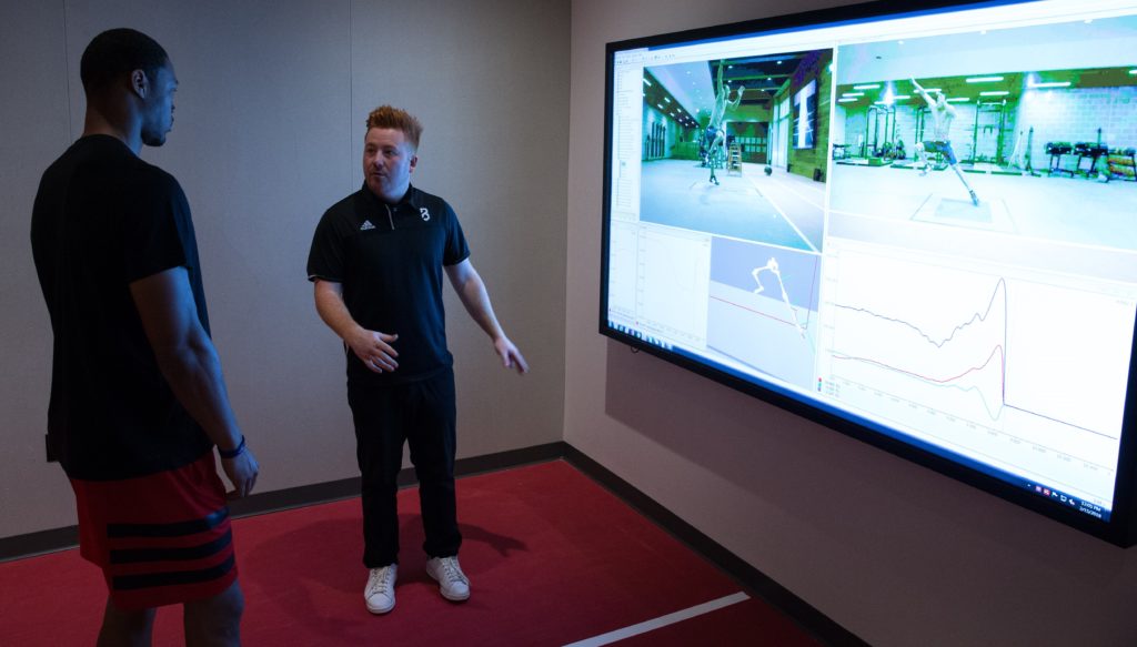 An athlete looks at a big screen projection of his workout and the data is produced while a coach talks with him.
