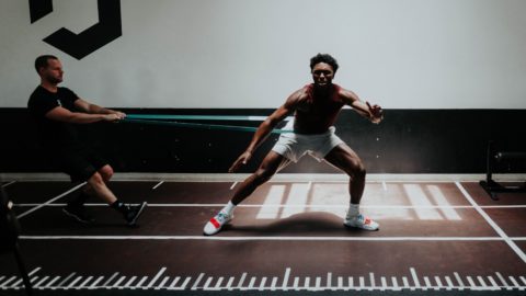 An athlete works on his lateral movement on an indoor track, wearing a stretching band around his waist, as a coach holds the other end of the band.