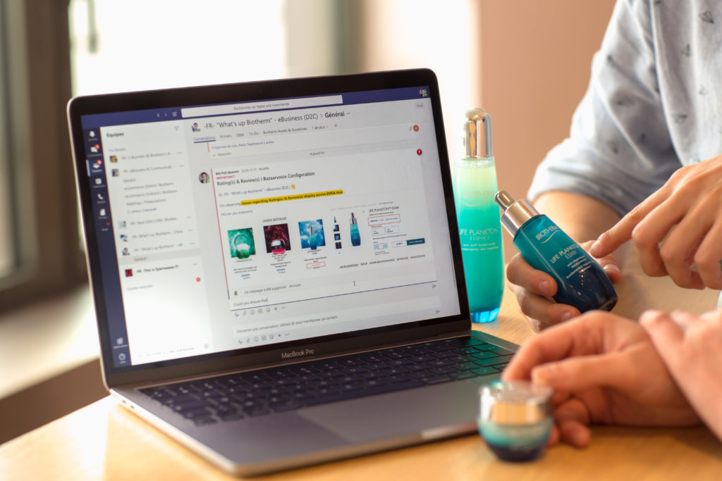 An open laptop displays conversations in Microsoft Teams as hands hold perfume bottles nearby. 
