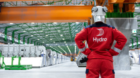 A worker is facing away from the camera and wearing a red Norsk Hydro jacket while looking out at a factory floor.