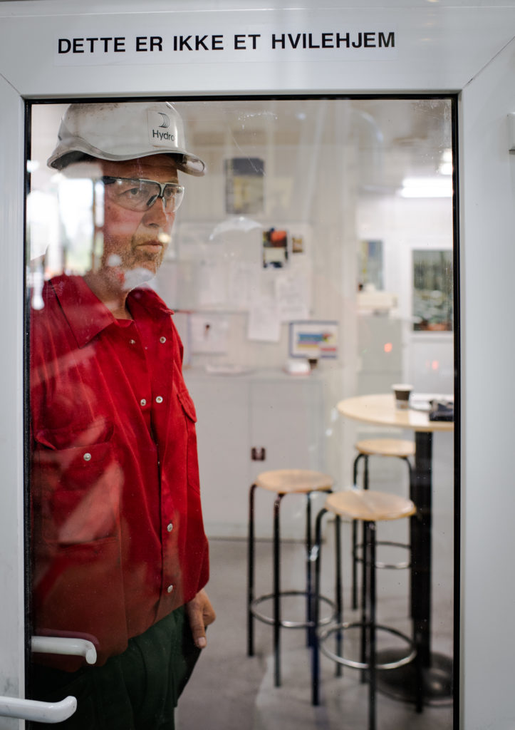 A Norsk Hydro employee in a hard hat looks out from behind a glass door inside a factory. 