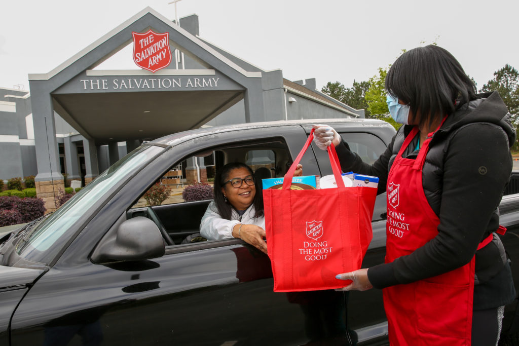 Woman in Salvation Army apron and mask handing off a bag of food to another woman in a car