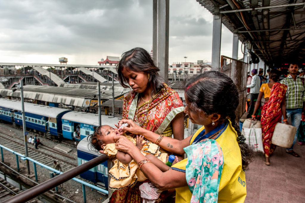 Photo of two young women vaccinating a baby outdoors at a train station in India.