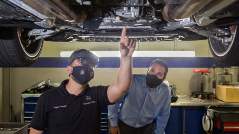 An auto technician in a HoloLens 2 looks up at the undercarriage of a car while pointing to a specific part while a second technician, to the right, looks at the same part. Both are wearing face masks.
