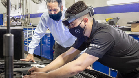 An auto repair technician wearing a HoloLens 2 and a face mask works with his hands on a Mercedes-Benz car engine while his supervisor, in a face mask, watches over his shoulder.