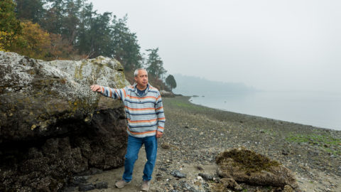 A man stands against a seashore boulder, with his right arm perched onto the boulder, looking out at the water.