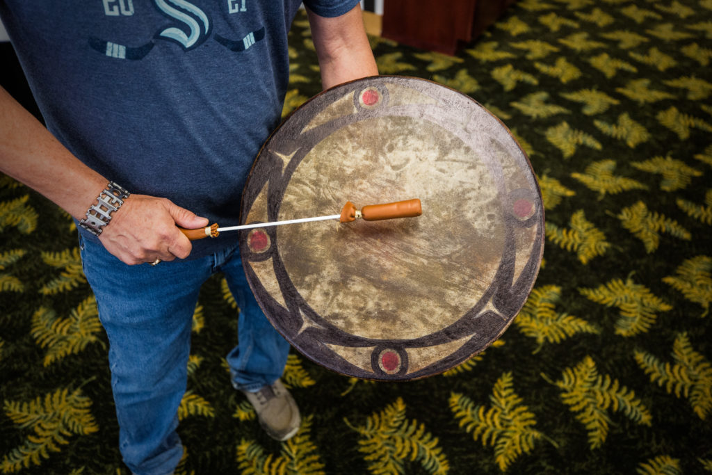 A Samish drum rests against a man's left hip as he places a drum stick on the surface. 