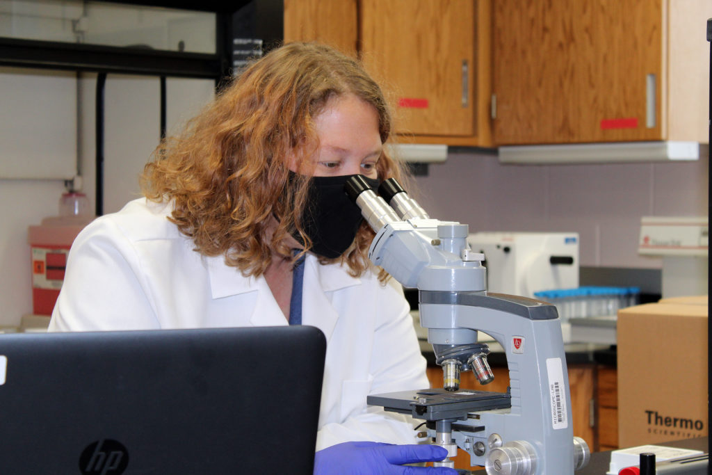A woman in a lab coat and a mask looks into a microscope. 