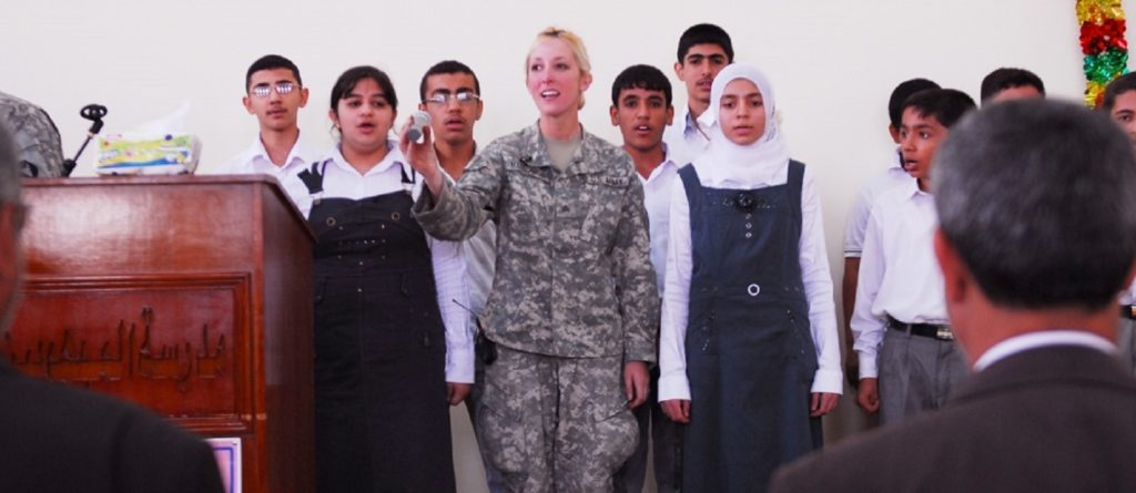 A U.S. soldier in Army fatigues stands on a stage flanked by three Iraqi people on each side, all of them signing together.