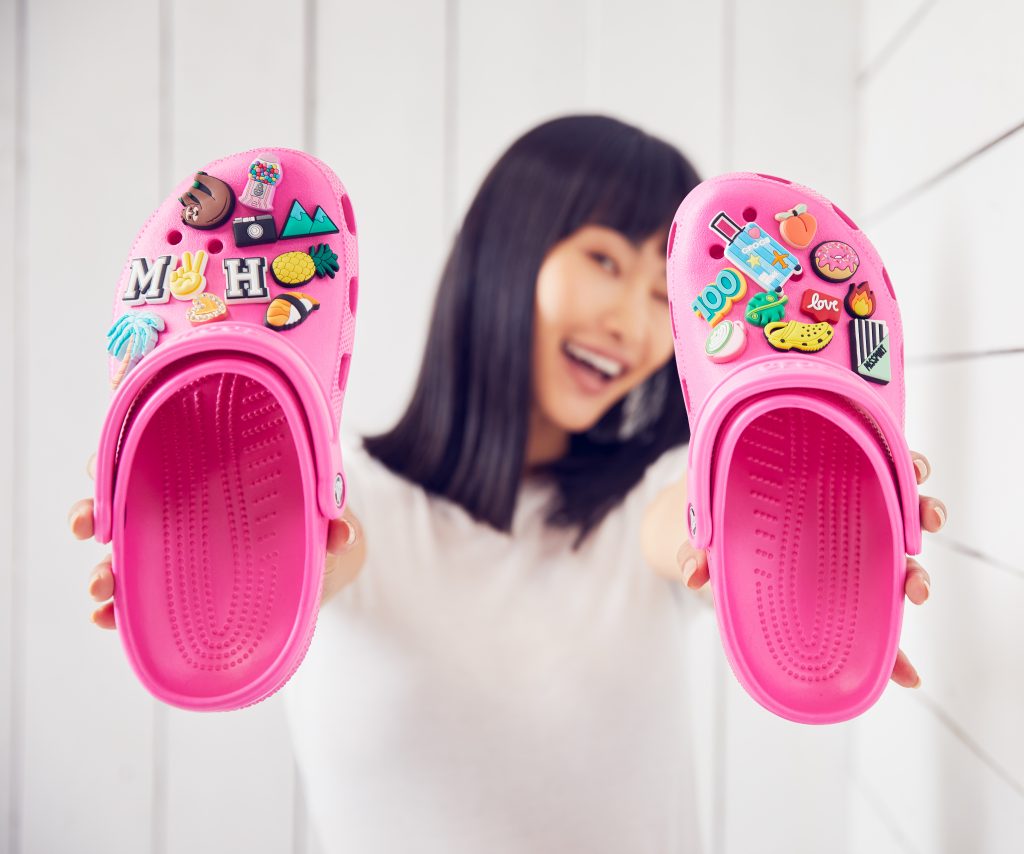 Smiling young woman slightly blurred in the background, standing against a white backdrop and holding up a pair of bright pink Crocs clogs adorned with charms, known as Jibbitz
