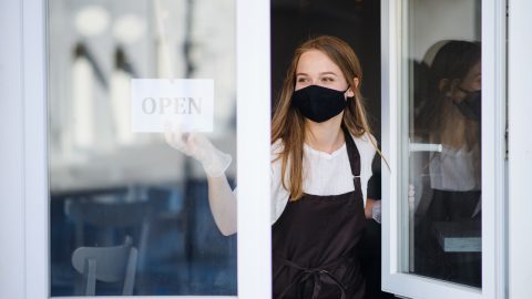 A woman wearing a face mask opens a cafe window.