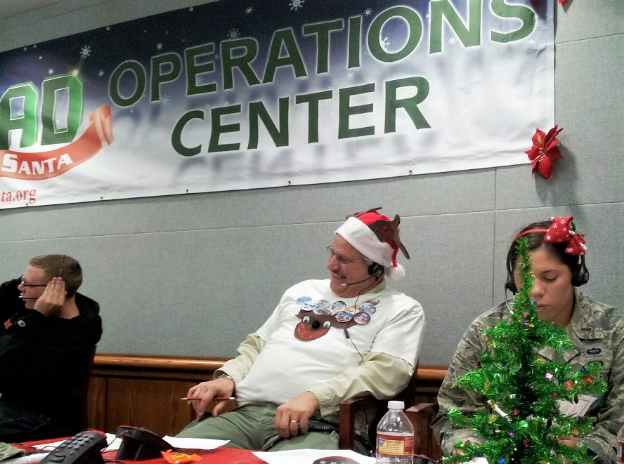 A man in a Santa hat speaks on his headset to a child calling the NORAD Santa Tracker on Christmas Eve.