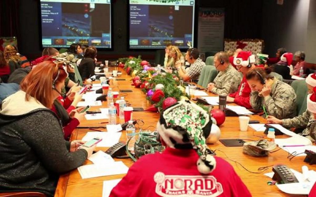 Several dozen people answer phone calls from kids on a recent Christmas Eve at the NORAD Tracks Santa operations center. Behind the rows of seated volunteers are two large wall screens showing a depiction of Santa and his reindeer in flight.