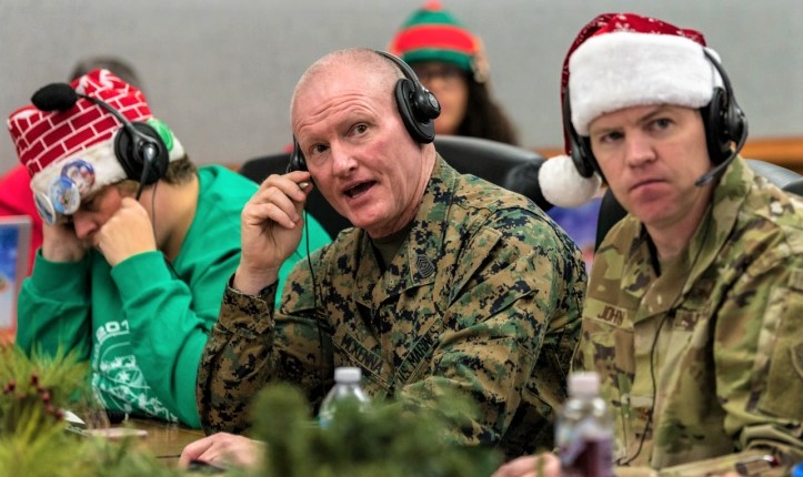 Two U.S. military members in camo uniforms are sitting next to each other at a table while wearing headsets. One of the soldiers is speaking to a caller. The second solider is wearing a Santa hat. 