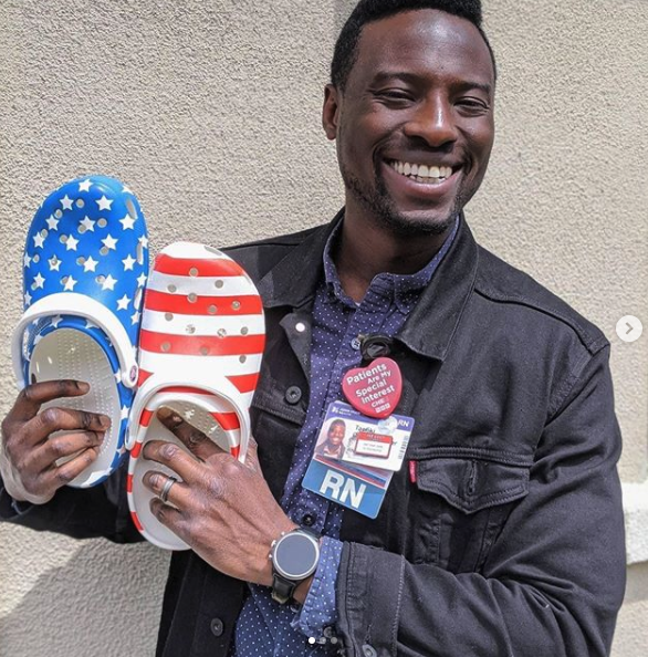 Smiling man wearing an RN badge holding up a pair of red, white and blue, stars and stripes-patterned Crocs clogs.