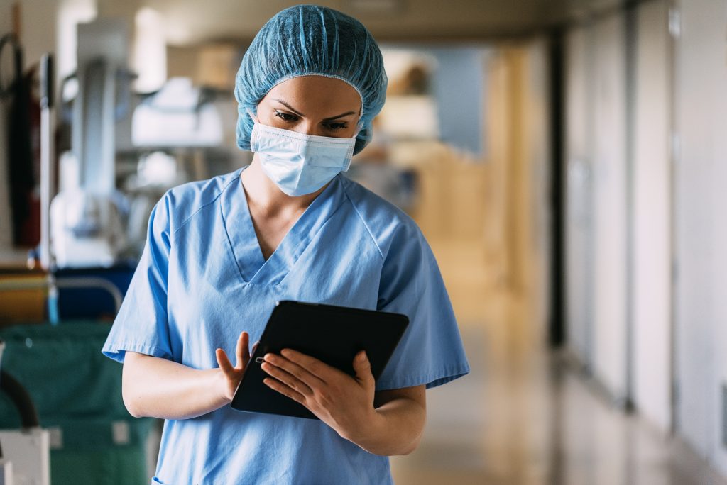 A nurse in a face mask and blue scrubs checks a smart tablet. 