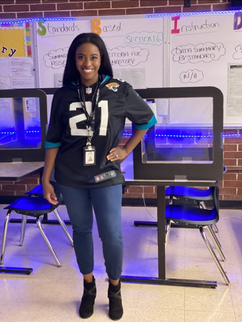 A woman wearing a football jersey stands in a school classroom with her left hand on her left hip.