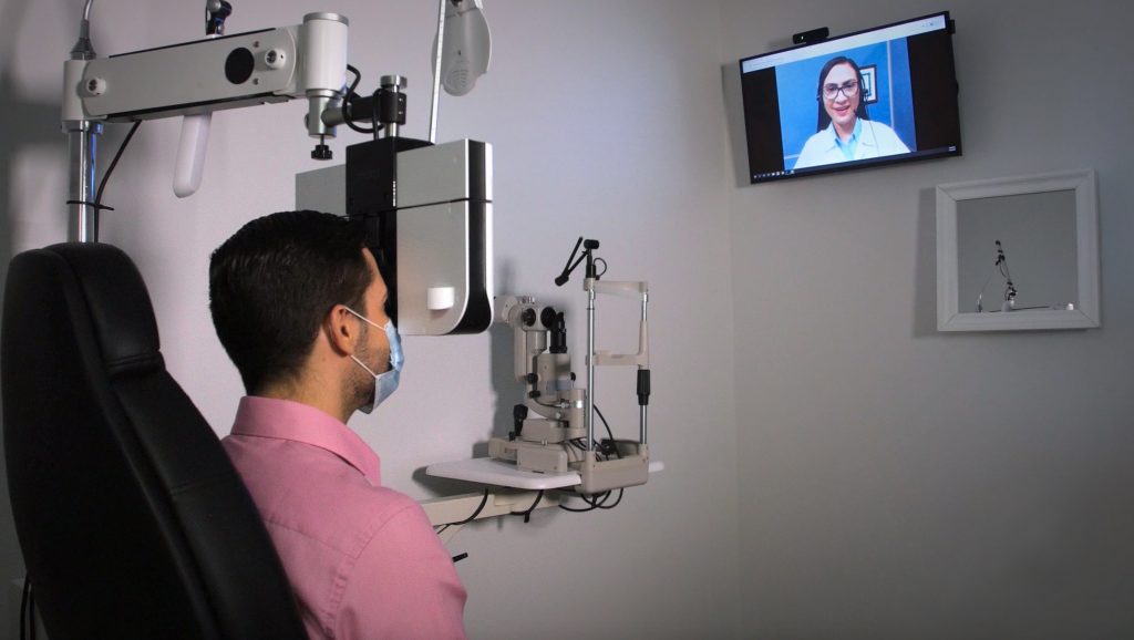 A patient looks into a refraction machine in an exam room while speaking with a remote eye doctor who appears on a screen attached to the wall.