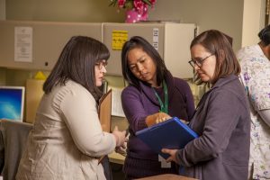 Photo of three female hospice employees in a medical setting, looking at a clipboard together.