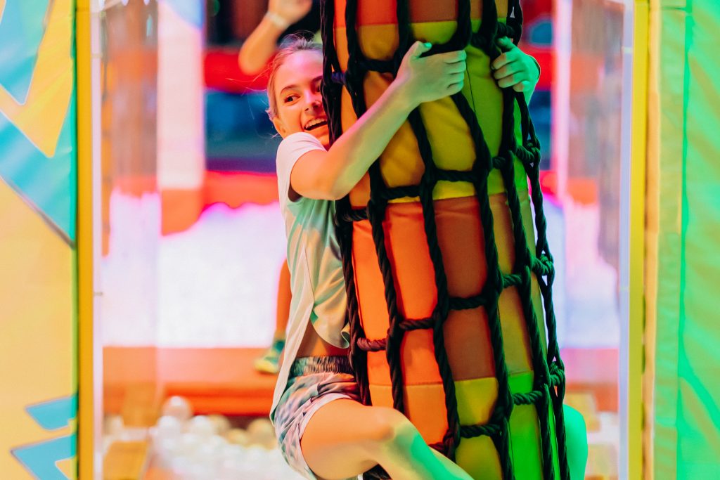 A girl smiles while hugging and hanging onto a vertical foam tube at an indoor amusement park.