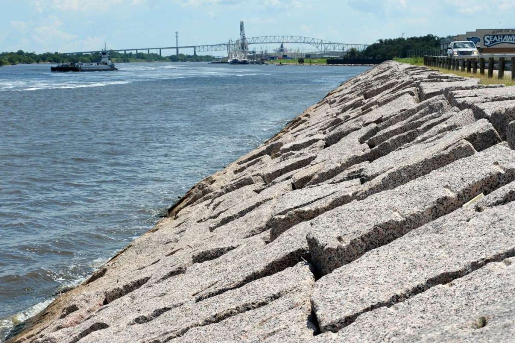 Photo showing a levee fortified with rocks on a river, with a boat and bridge in the background.