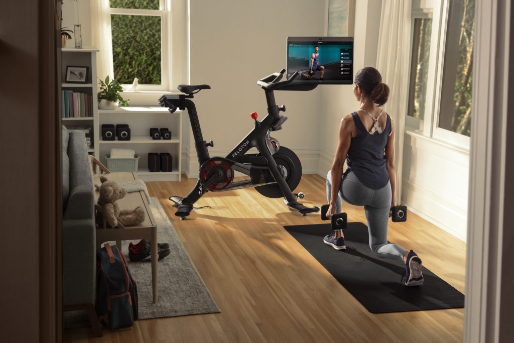 Photo of woman at home, doing lunges with weights beside a Peloton Bike. lunging with dumbell wei