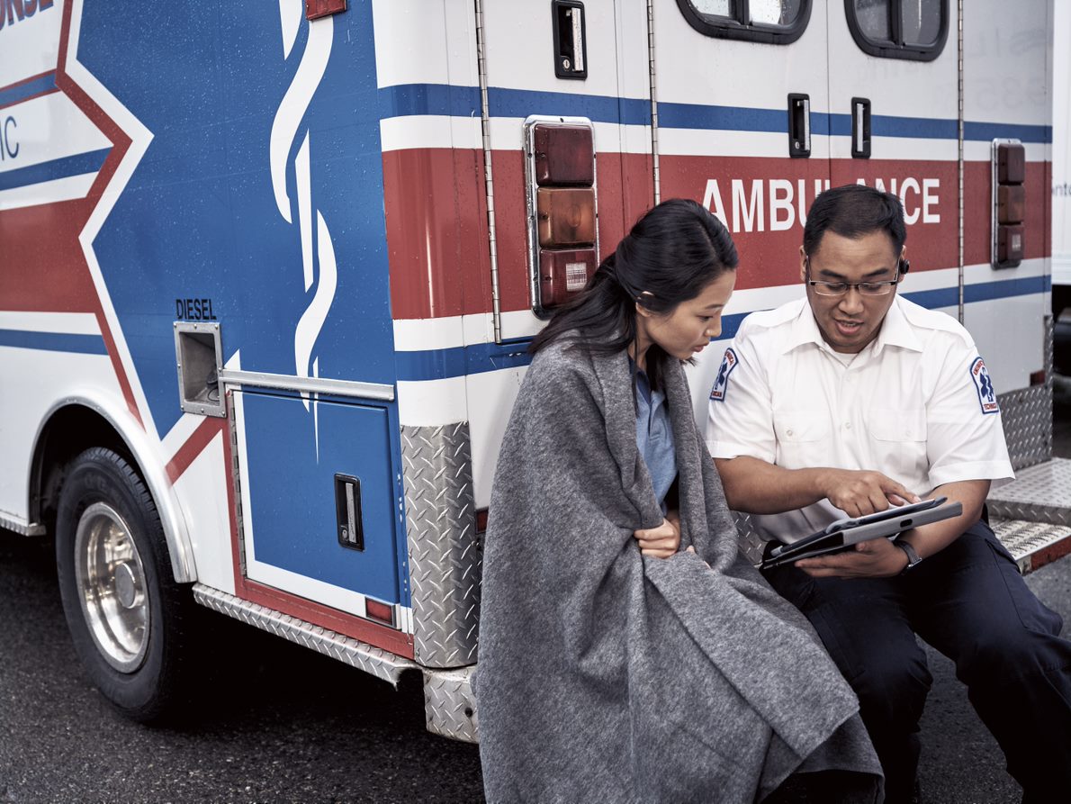 A woman sits on the bumper of an ambulance with an EMT