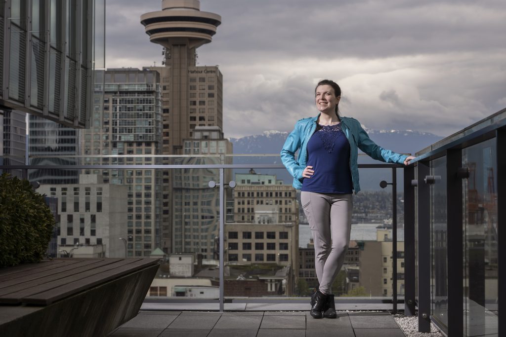 Photo of Katie Peters, staff developer for TELUS, standing on a deck at the company’s Vancouver, B.C. headquarters, with skyline and mountains in background.