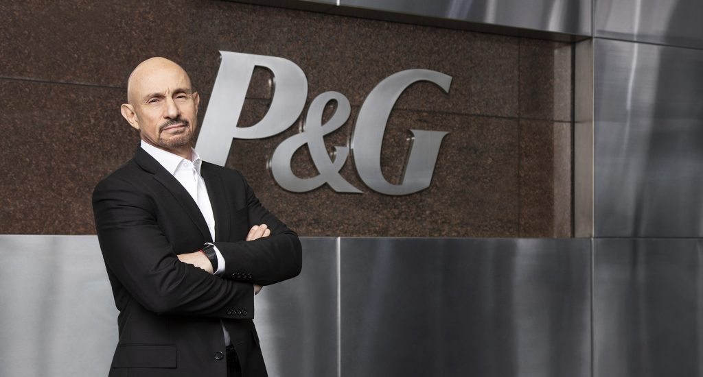 Vittorio Cretella stands with arms folded in front of a wall that shows the P&G logo.