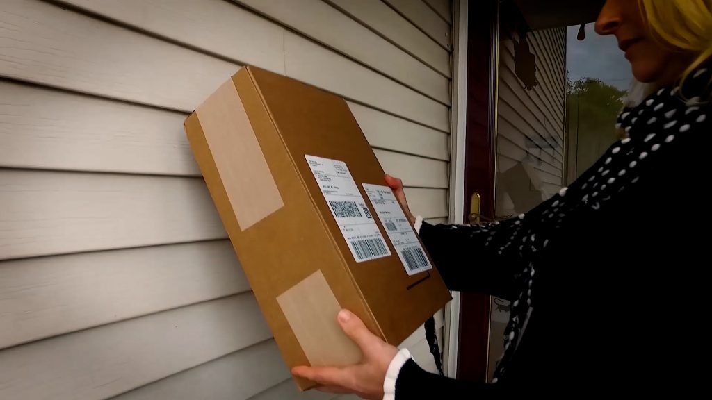 A woman holds and examines a shipping box from Wolverine Worldwide while standing on her porch. 
