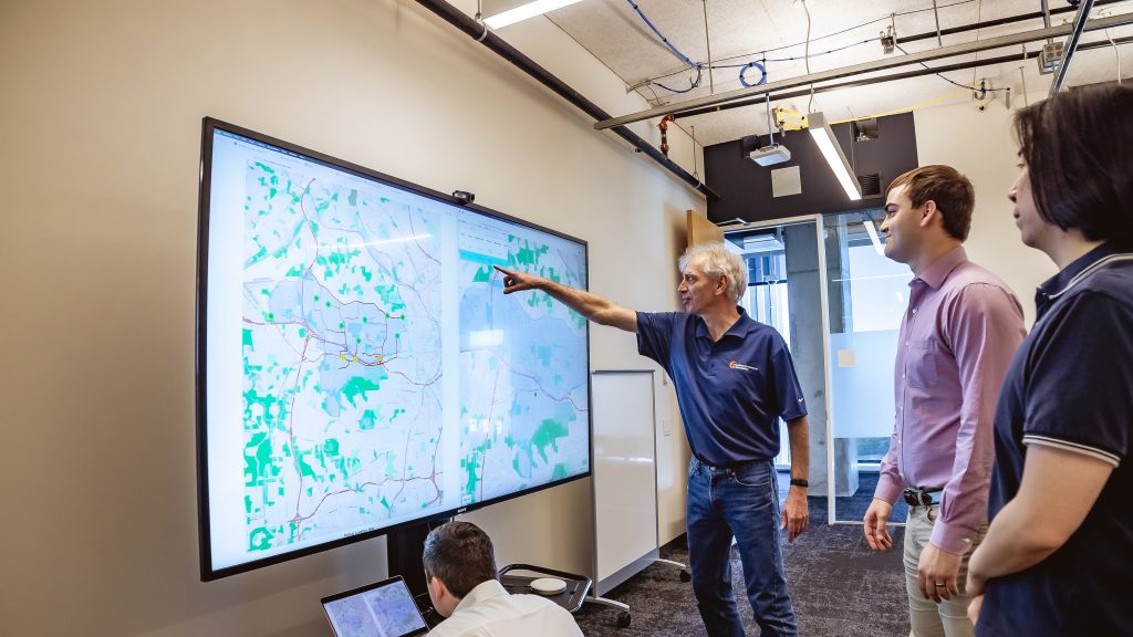 Georgia Tech professor Pascal Van Hentenryck, surrounded by three Ph.D. students, points to a large screen displaying the operations app for the MARTA Reach on-demand ride-share service in Atlanta. 