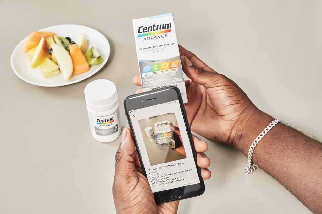 Close-up of hands holding a box of Centrum in one hand and a phone displaying the Seeing AI app in the other.