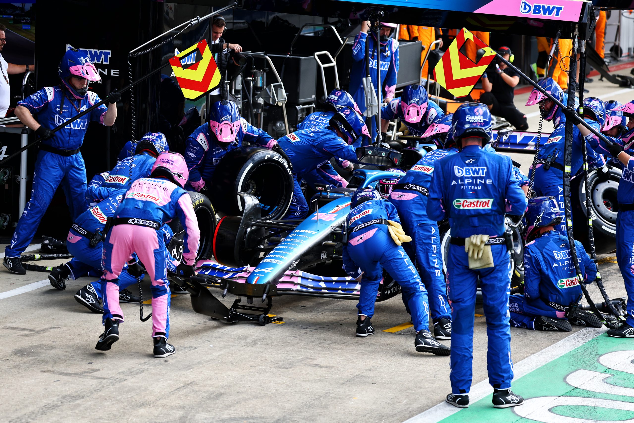 Alpine pit crew members in blue jumpsuits attend to Fernando Alonso's car during a pit stop, including changing the car's tires. 