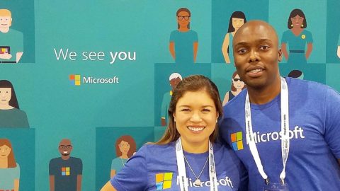 Two Microsoft employee smile in front of a backdrop that says We see you