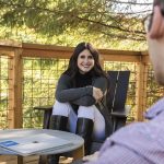 Smiling female employee sitting in an Adirondack chair on an outside deck talking to a colleague