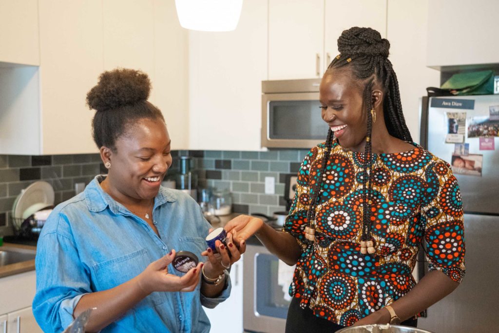 Two women in a kitchen laughing as they hold a jar of shea butter