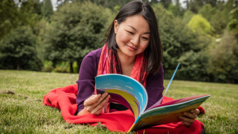 A photo of a woman lying on a blanket in the grass reading a book