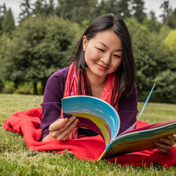 A photo of a woman lying on a blanket in the grass reading a book