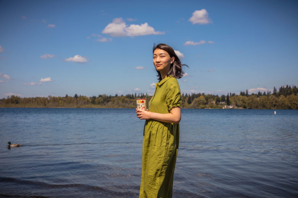A woman in a green dress who is holding a coffee cup and standing at the shore of a lake